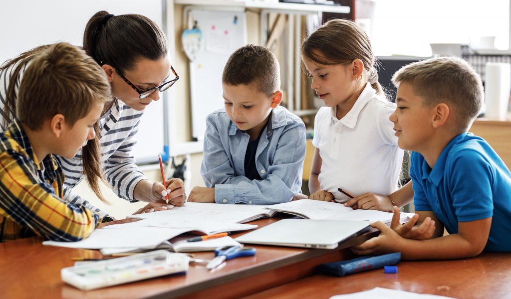 Female teacher helps school kids to finish they lesson.They sitting all together at one desk.